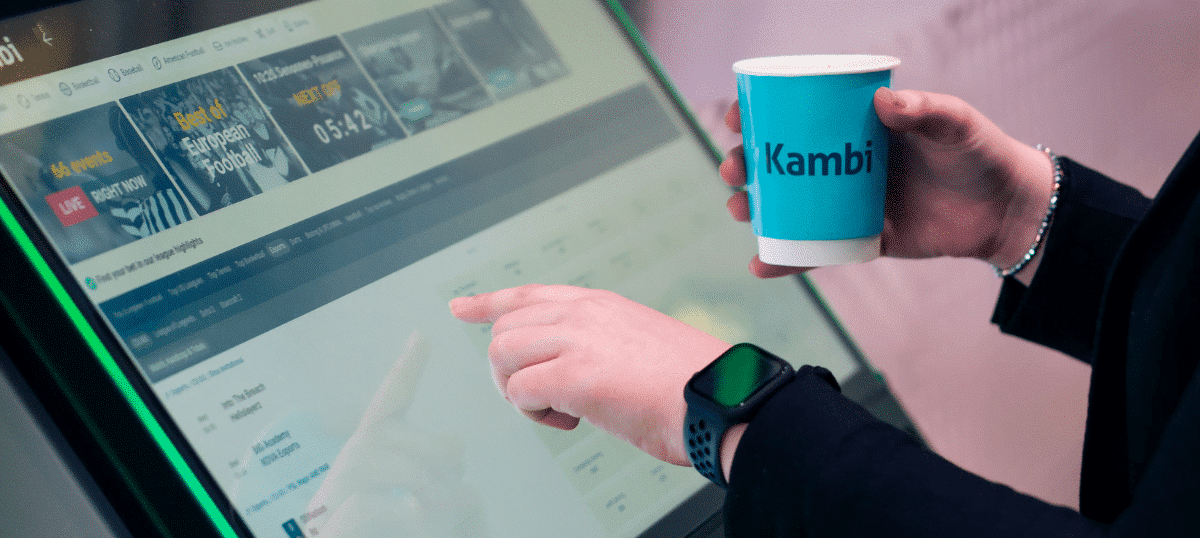 A person using a Kambi betting machine while holding a Kambi-branded paper cup.