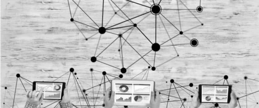 A black and white image of Hhands holding tablets displaying data graphs, connected by intersecting lines and nodes, against a textured wooden background.