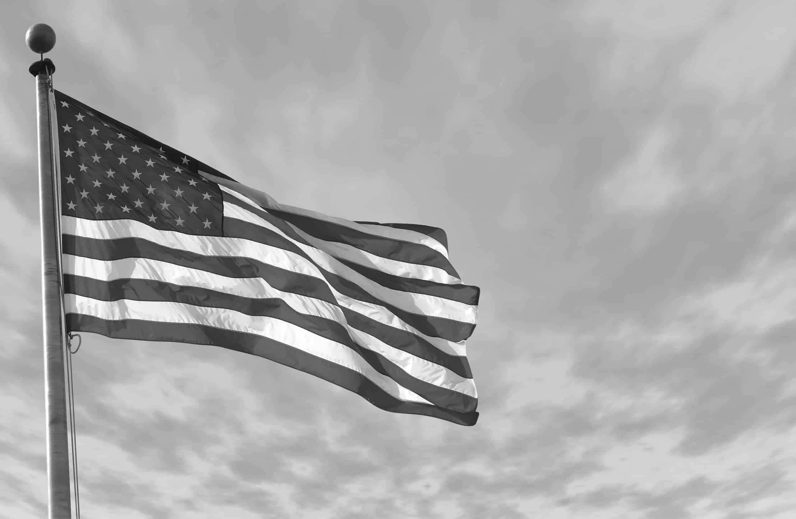 A black and white close up picture of a American flag outside moving in the wind.